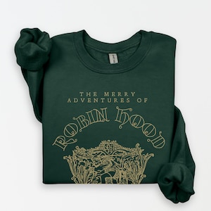 May include: A dark green sweatshirt with the text "THE MERRY ADVENTURES OF ROBIN HOOD" in a gold-colored, decorative font. The design includes an illustration of Robin Hood and his forest setting. The sweatshirt is folded, showing the rolled-up sleeves.