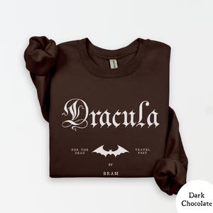 Op de afbeelding: Donkerbruin sweatshirt met het woord "Dracula" in een gotisch lettertype. Daaronder staat een vleermuisafbeelding en de zinnen "FOR THE DEAD" en "TRAVEL FAST". De tekst "BY BRAM" is ook opgenomen.