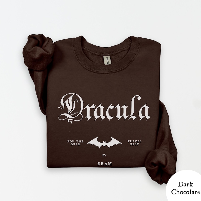 Op de afbeelding: Donkerbruin sweatshirt met het woord "Dracula" in een gotisch lettertype. Daaronder staat een vleermuisafbeelding en de zinnen "FOR THE DEAD" en "TRAVEL FAST". De tekst "BY BRAM" is ook opgenomen.
