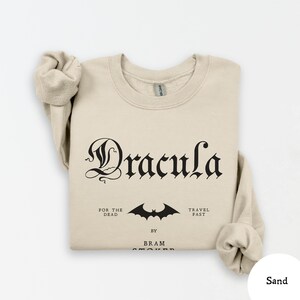 Op de afbeelding: Zandkleurige sweatshirt met het woord "Dracula" in een gotisch lettertype. Daaronder een vleermuisafbeelding en de zinnen "For the Dead" en "Travel Fast". De naam van de auteur, "Bram Stoker", staat er ook op.