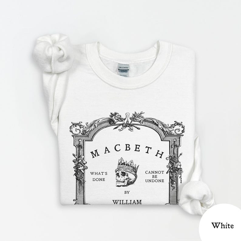 May include: White sweatshirt featuring a black and white graphic design. The design includes the word "Macbeth" above a skull wearing a crown, with text that reads "What's Done Cannot Be Undone" and "By William."