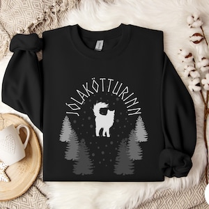 May include: Black sweatshirt with a white graphic featuring a cat, pine trees, and the text "JÓLAKÖTTURINN" in a semi-circle. The design is centered on the chest. The sweatshirt has long sleeves and a crew neck.
