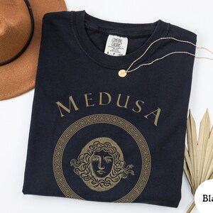 May include: Black t-shirt with a gold Medusa graphic. The design features the word "MEDUSA" above a circular illustration of Medusa's face, surrounded by a Greek key border. A gold necklace and a brown hat are also in the image.