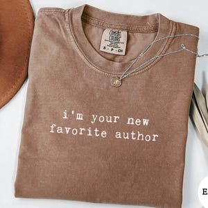 May include: Espresso-colored t-shirt with the text "I'm your new favorite author" in white. A silver necklace with a pendant is draped on the shirt. A brown hat and dried leaves are in the background.