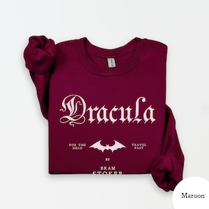 Op de afbeelding: Bordeaux sweatshirt met het woord "Dracula" in een gotisch lettertype. Onder de titel staat een vleermuisafbeelding en de zinnen "For the Dead" en "Travel Fast". De naam van de auteur, Bram Stoker, staat ook op de trui.