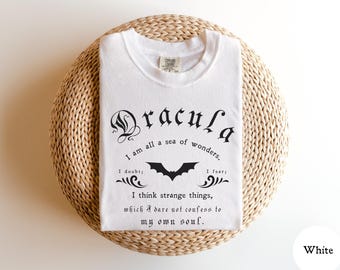 May include: White t-shirt with the word "Dracula" in gothic font, a bat graphic, and a quote. The shirt is folded on a woven, circular surface. The text reads "I am all a sea of wonders, I doubt; I fear; I think strange things, which I dare not confess to my own soul."