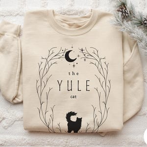 May include: A sand-colored crewneck sweatshirt featuring a black graphic print. The design includes a crescent moon, stars, and a circular branch frame surrounding the text 'the YULE cat' above a silhouette of a fluffy cat with a bushy tail.