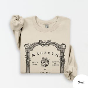 May include: Sand-colored sweatshirt with the text "Macbeth" in a decorative frame, featuring a skull wearing a crown. The text "What's Done Cannot Be Undone" is also displayed. The sweatshirt is a Gildan brand.