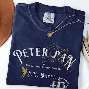 May include: True navy blue t-shirt with the text "PETER PAN" and "The Boy Who Wouldn't Grow Up" in white and gold. A gold necklace with a pendant rests on the shirt. The shirt also features a fairy illustration.