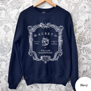 May include: Navy blue sweatshirt featuring a white graphic design from Shakespeare's play, Macbeth. The design includes the title, a skull wearing a crown, and the phrases "What's Done" and "Cannot Be Undone". The text "By William Shakespeare" is also included.