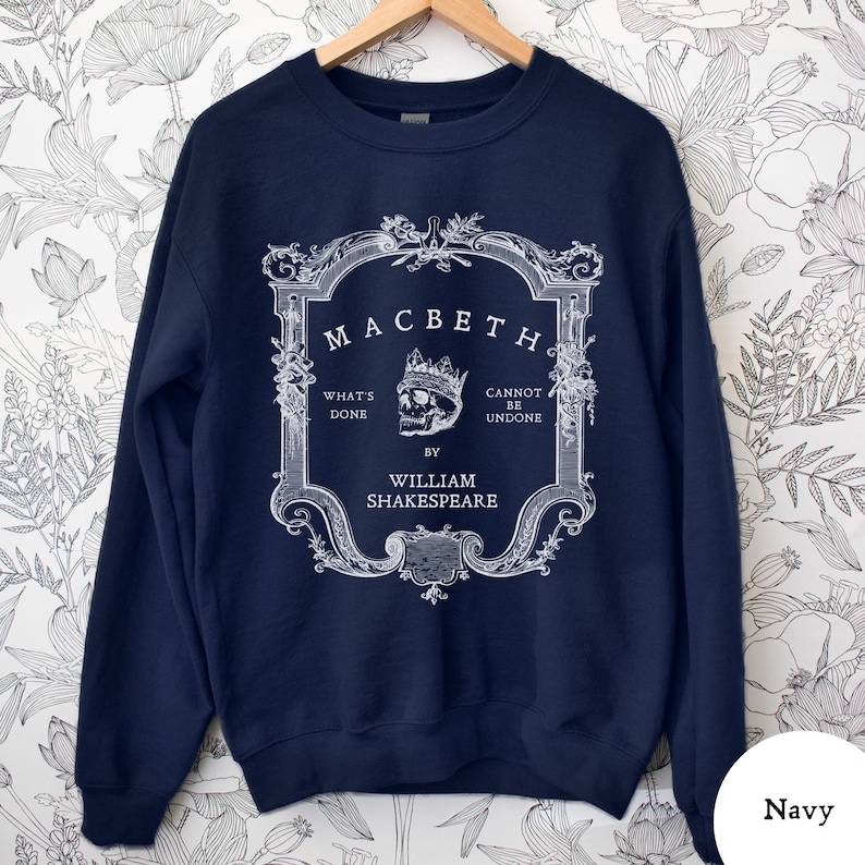 May include: Navy blue sweatshirt featuring a white graphic design from Shakespeare's play, Macbeth. The design includes the title, a skull wearing a crown, and the phrases "What's Done" and "Cannot Be Undone". The text "By William Shakespeare" is also included.