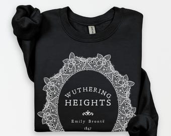 May include: A black crewneck sweatshirt featuring a white graphic print of an oval lace frame. Inside the frame is the text WUTHERING HEIGHTS, Emily Brontë, and 1847. The sweatshirt has long sleeves and a ribbed collar with a visible brand tag.