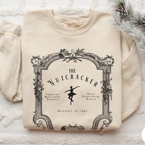 May include: A sand-colored crewneck sweatshirt featuring a black graphic print of an ornate frame. Inside the frame is the text THE NUTCRACKER, Imperial Mariinsky Theatre, Saint Petersburg, Russia, and December 18, 1891, with a ballerina silhouette.