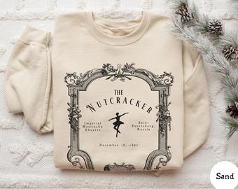 May include: A sand-colored crewneck sweatshirt featuring a black graphic print of an ornate frame. Inside the frame is the text THE NUTCRACKER, Imperial Mariinsky Theatre, Saint Petersburg, Russia, and December 18, 1891, with a ballerina silhouette.