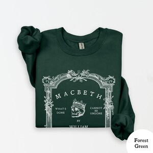 May include: Forest green sweatshirt with the word "Macbeth" in a decorative frame. The design includes a skull wearing a crown, and the phrases "What's Done" and "Cannot Be Undone" are also visible. The text "By William" is at the bottom.