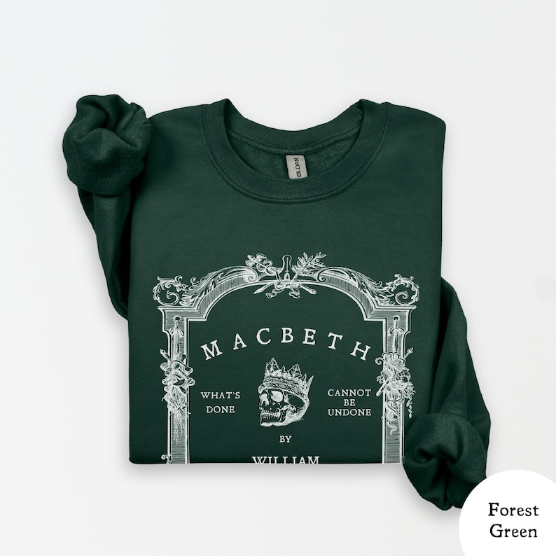 May include: Forest green sweatshirt with the word "Macbeth" in a decorative frame. The design includes a skull wearing a crown, and the phrases "What's Done" and "Cannot Be Undone" are also visible. The text "By William" is at the bottom.