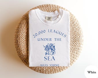 May include: A white t-shirt featuring blue text that reads 20,000 LEAGUES UNDER THE SEA and JULES VERNE, with a blue graphic illustration of an octopus centered between the words. The shirt has a crew neckline and a Comfort Colors brand label.