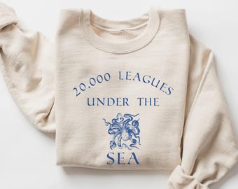 May include: A sand-colored crewneck sweatshirt featuring blue text that reads 20,000 LEAGUES UNDER THE SEA, accompanied by a detailed blue graphic illustration of an octopus positioned between the words UNDER THE and SEA.