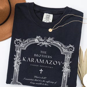 May include: Black t-shirt featuring the text "The Brothers Karamazov" by Fyodor Dostoevsky, with a decorative frame and cross design. A gold necklace with a small pendant rests on the shirt. A brown hat is visible in the background.
