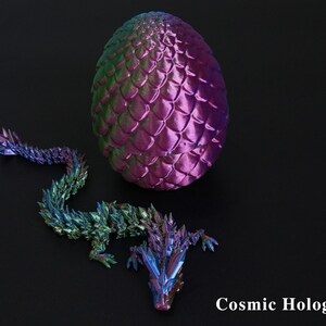 Pu&ograve; includere: Un uovo di drago olografico e una statuina di drago abbinata. L'uovo &egrave; viola e verde con una trama a scaglie. Il drago &egrave; multicolore con un aspetto cristallino. Il testo "Cosmic Hologram" &egrave; visibile.