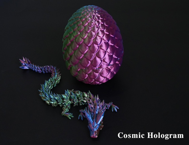 Pu&ograve; includere: Un uovo di drago olografico e una statuina di drago abbinata. L'uovo &egrave; viola e verde con una trama a scaglie. Il drago &egrave; multicolore con un aspetto cristallino. Il testo "Cosmic Hologram" &egrave; visibile.