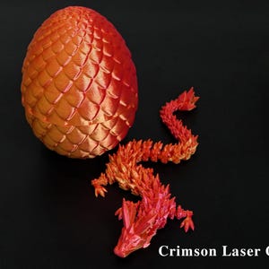 Pu&ograve; includere: Un uovo di drago e una statuina di drago stampati in 3D, nei toni del rosso e dell'arancione. L'uovo presenta un motivo strutturato a forma di scaglie. Il drago &egrave; posato con il corpo curvo. Il testo "Crimson Laser Glow" &egrave; in basso.