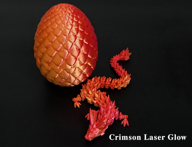 Pu&ograve; includere: Un uovo di drago e una statuina di drago stampati in 3D, nei toni del rosso e dell'arancione. L'uovo presenta un motivo strutturato a forma di scaglie. Il drago &egrave; posato con il corpo curvo. Il testo "Crimson Laser Glow" &egrave; in basso.