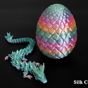 Pu&ograve; includere: Un uovo di drago decorativo e una statuina abbinata. L'uovo presenta un mix di rosa, arancione, verde e blu, con un motivo a scaglie. Il drago &egrave; turchese con dettagli cristallini. Il testo "Silk Candy" &egrave; visibile.