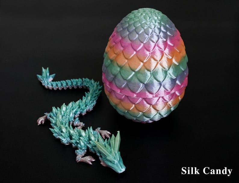 Pu&ograve; includere: Un uovo di drago decorativo e una statuina abbinata. L'uovo presenta un mix di rosa, arancione, verde e blu, con un motivo a scaglie. Il drago &egrave; turchese con dettagli cristallini. Il testo "Silk Candy" &egrave; visibile.