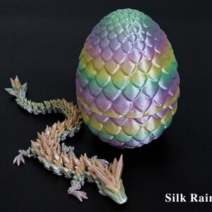 Pu&ograve; includere: Un uovo di drago multicolore stampato in 3D e una statuina di drago abbinata. L'uovo presenta una vivace miscela di colori arcobaleno, con un motivo strutturato a forma di scaglia. Il drago &egrave; nelle tonalit&agrave; del verde, beige e rosa. Il testo "Silk Rainbow" &egrave; visibile.