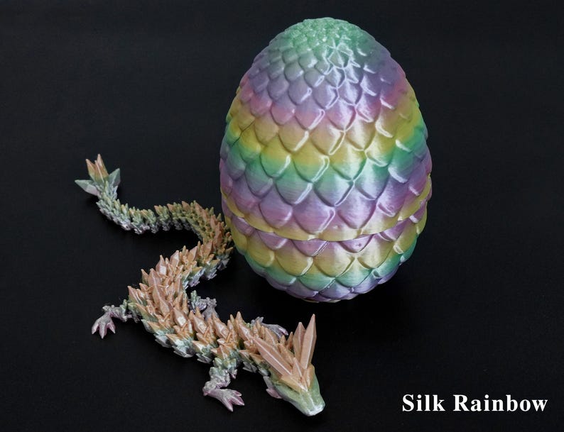 Pu&ograve; includere: Un uovo di drago multicolore stampato in 3D e una statuina di drago abbinata. L'uovo presenta una vivace miscela di colori arcobaleno, con un motivo strutturato a forma di scaglia. Il drago &egrave; nelle tonalit&agrave; del verde, beige e rosa. Il testo "Silk Rainbow" &egrave; visibile.