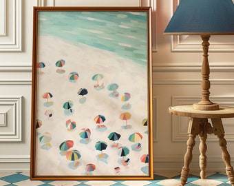 May include: A framed art print depicts a beach scene with colorful umbrellas scattered across the sand. The artwork has a gold-colored frame and is leaning against a white wall. A blue lampshade sits atop a wooden lamp.