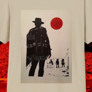 May include: Beige t-shirt featuring a graphic print of a figure in a poncho and hat, with a red sun in the background. The design is a silhouette style, with a western theme. The shirt is made of a soft material.