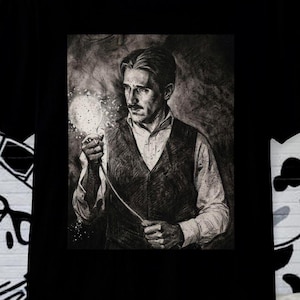 May include: Black t-shirt featuring a grayscale portrait of Nikola Tesla holding a glowing light bulb. The image is centered on the shirt, showcasing the inventor's likeness. The shirt's design is a tribute to the famous scientist.
