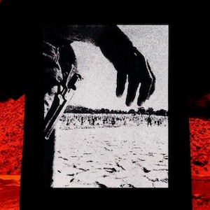May include: Black t-shirt featuring a black and white graphic of a hand holding a gun, with a crowd in the background. The shirt has a crew neck and short sleeves. The background is a fiery red and orange.