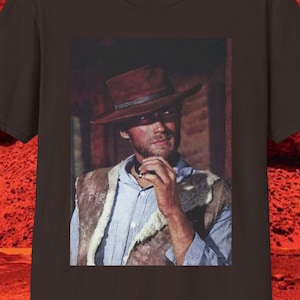 May include: A dark brown t-shirt featuring a framed image of a man in a brown hat and vest. The man is holding a cigarette. The background is a red desert landscape.