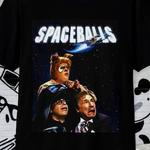 May include: Black t-shirt featuring the movie title "SPACEBALLS" above a graphic of three characters from the film. The graphic is set against a starry background. The t-shirt is made of a soft material.