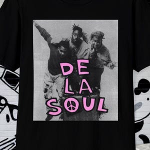 以下が含まれることがあります： 黒いTシャツ。ピンク色のグラフィックで3人の人物と「DE LA SOUL」の文字が描かれています。デザインには平和のシンボルが含まれています。Tシャツは落書きアートの背景に表示されています。