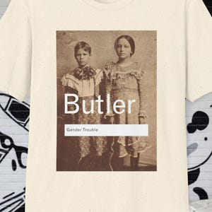 May include: Cream-colored t-shirt featuring a sepia-toned image of two children in vintage clothing. The image includes the text "Butler" above "Gender Trouble" in a white rectangle. The shirt is displayed against a backdrop of graffiti art.