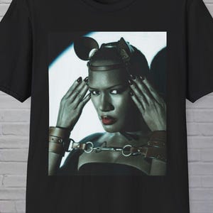 May include: Black t-shirt with a portrait of a person with dark hair, red lipstick, and leather accessories. The image is a close-up, with the person's hands near their head. The shirt is made of a soft material.