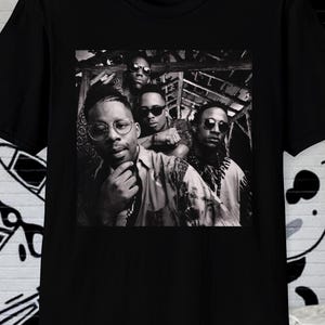 以下が含まれることがあります： 黒いTシャツ。白黒写真で、グループの人々が写っています。Tシャツの画像には、サングラスをかけてカメラを見ている5人が写っています。Tシャツは半袖で、クルーネックです。