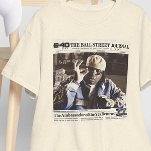 May include: Cream-colored t-shirt featuring a newspaper-style graphic with the text "E-40 THE BALL STREET JOURNAL" and an image of a person. The text "The Ambassador of the Yay Returns!" is also visible. A white baseball cap hangs on a wooden chair.
