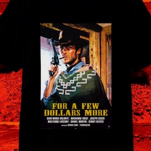 May include: Black t-shirt featuring a movie poster for "For a Few Dollars More." The poster depicts a man in a cowboy hat holding a gun. The shirt includes the movie title and cast names.