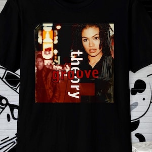 May include: Black t-shirt featuring a graphic of a woman with the words "groove theory" in red and white. The image on the shirt has a retro aesthetic, with a blurred background of city lights.