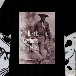 May include: Black t-shirt featuring a sepia-toned image of a Black cowboy with the text "Black Cowboys of Texas." The cowboy is wearing a hat, scarf, and holding a bag. The shirt is a classic crew neck.