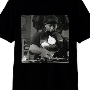 May include: Black t-shirt featuring a black and white image of a person holding a vinyl record. The image shows a DJ at work, with a turntable and the record in focus. The shirt is a classic crew neck style.