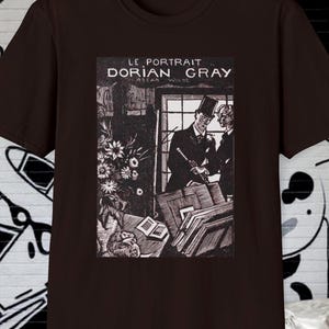 May include: A dark brown t-shirt featuring a black and white illustration from "Le Portrait de Dorian Gray." The graphic depicts two men in formal attire, surrounded by books and flowers. The text "LE PORTRAIT DORIAN GRAY" is printed above the illustration.