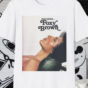 Op de afbeelding: Wit T-shirt met de filmposter van "Foxy Brown" met Pam Grier. De afbeelding toont een vrouw met gesloten ogen. De tekst "Pam Grier... Foxy Brown" staat boven de afbeelding.