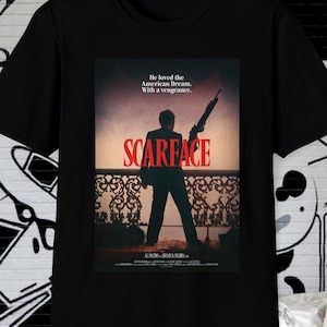 May include: Black t-shirt with a movie poster design. The poster shows a silhouette of a man holding a gun, with the word "SCARFACE" in red. The text "He loved the American Dream. With a vengeance." is also visible.