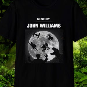 Puede incluir: Camiseta negra con un gráfico de una luna llena y siluetas de personajes de películas. El texto "MUSIC BY JOHN WILLIAMS" está impreso encima del gráfico. Un diseño único para los cinéfilos.